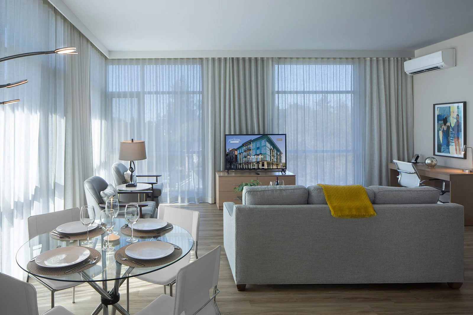 A modern apartment-style suite featuring a cozy living area with a gray sofa, a glass dining table set for four, a flat-screen TV, and a desk. Natural light filters through sheer curtains, enhancing the spacious design.