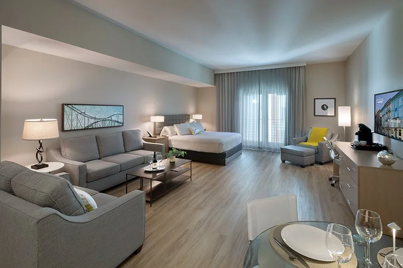 Spacious and modern studio suite featuring a comfortable bed, seating area with a sofa, and a dining table, with large windows allowing natural light.